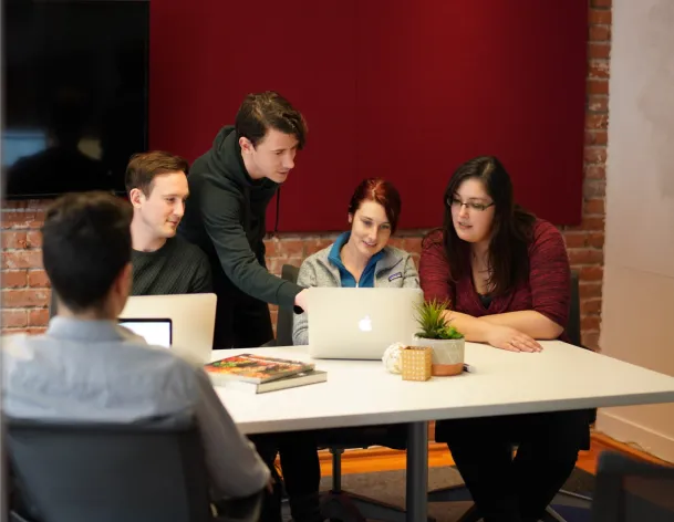 Pixieset team gathered around a laptop during a collaborative team meeting inside a creative office with brick walls and red acoustic panels.